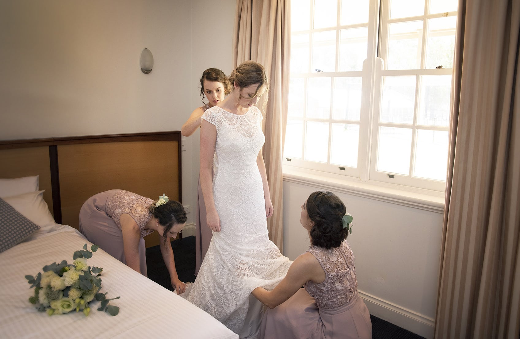 serafino wines wedding - claire & arron 26 Bridesmaids helping put on wedding dress next to window