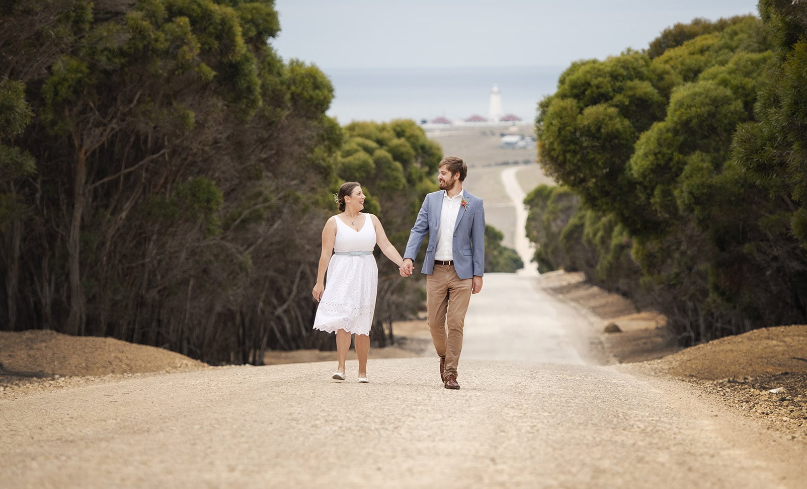 kangaroo island elopement - claire & jacob 17 Claire and Jacob walking hand in hand along a tree-lined gravel road with Cape Willoughby Lighthouse in the distance