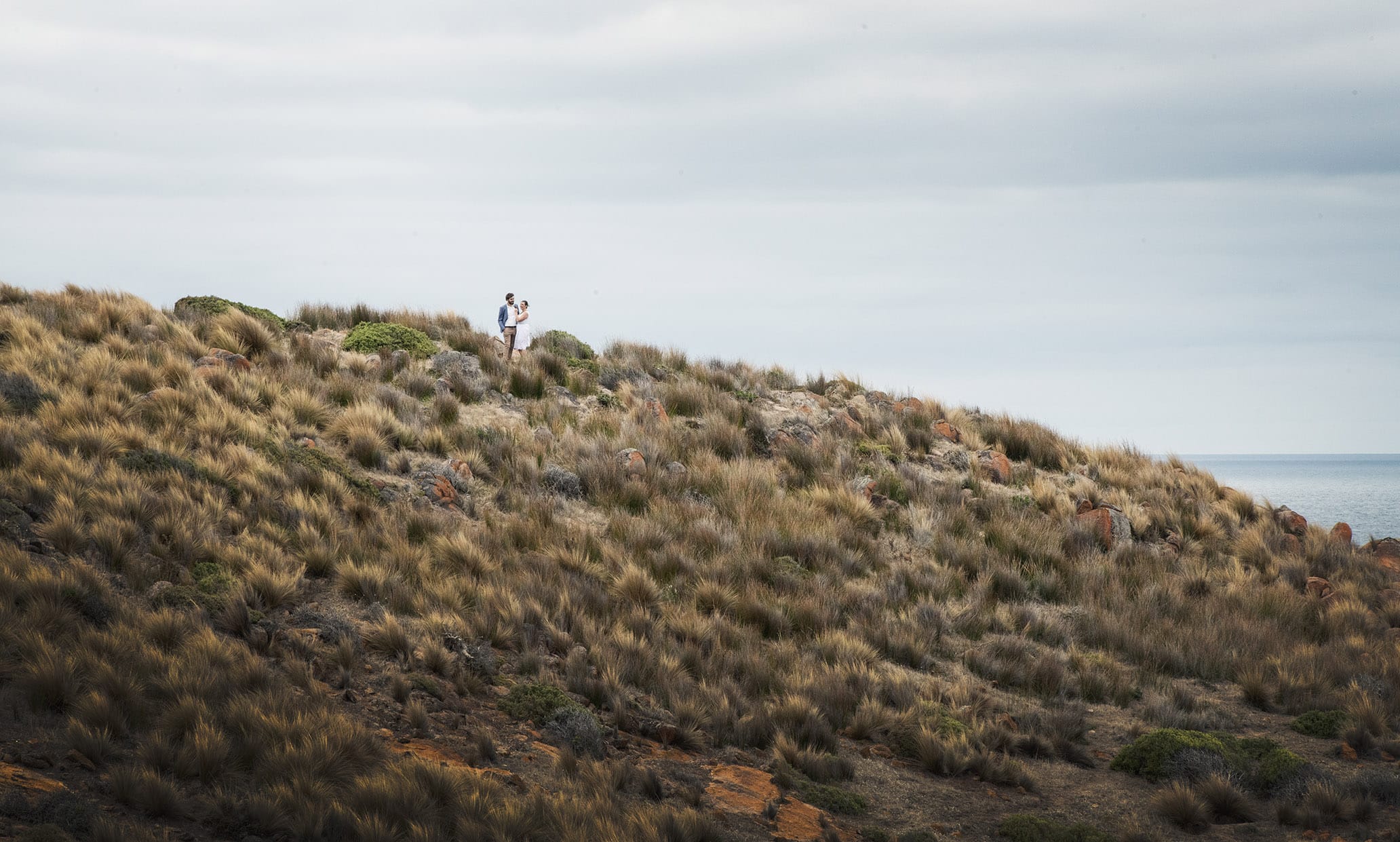 kangaroo island elopement - claire & jacob 18 Claire and Jacob embracing on a grassy, rocky coastal hill overlooking the sea on Kangaroo Island