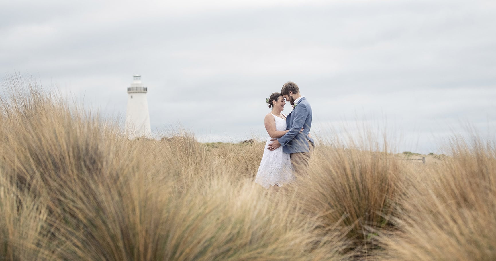 kangaroo island elopement - claire & jacob 22 Claire and Jacob embracing in coastal grass with Cape Willoughby Lighthouse behind them on Kangaroo Island