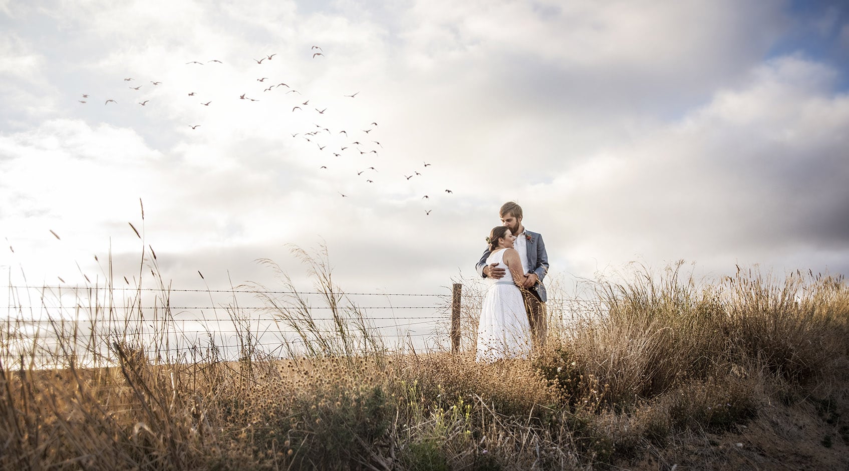 kangaroo island elopement - claire & jacob 12 Claire and Jacob embracing beside a wire fence in coastal grassland as birds fly overhead on Kangaroo Island