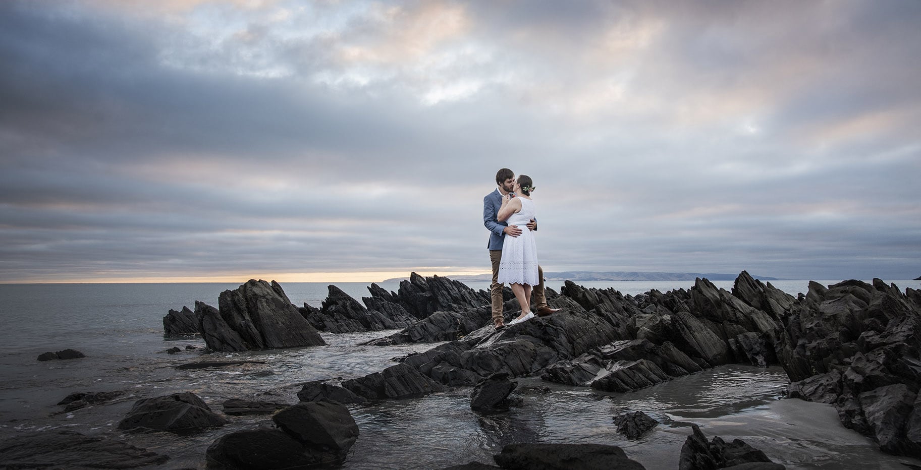 kangaroo island elopement - claire & jacob 6 Claire and Jacob embracing on dark coastal rocks at Penneshaw beach beneath an overcast sunrise sky