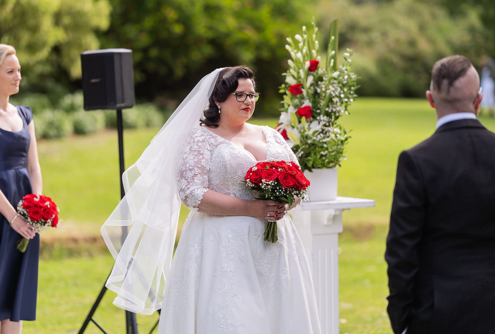 botanic gardens palm house wedding - kelly & david 94 Bride in a lace wedding dress and veil holding a red rose bouquet during an outdoor garden ceremony