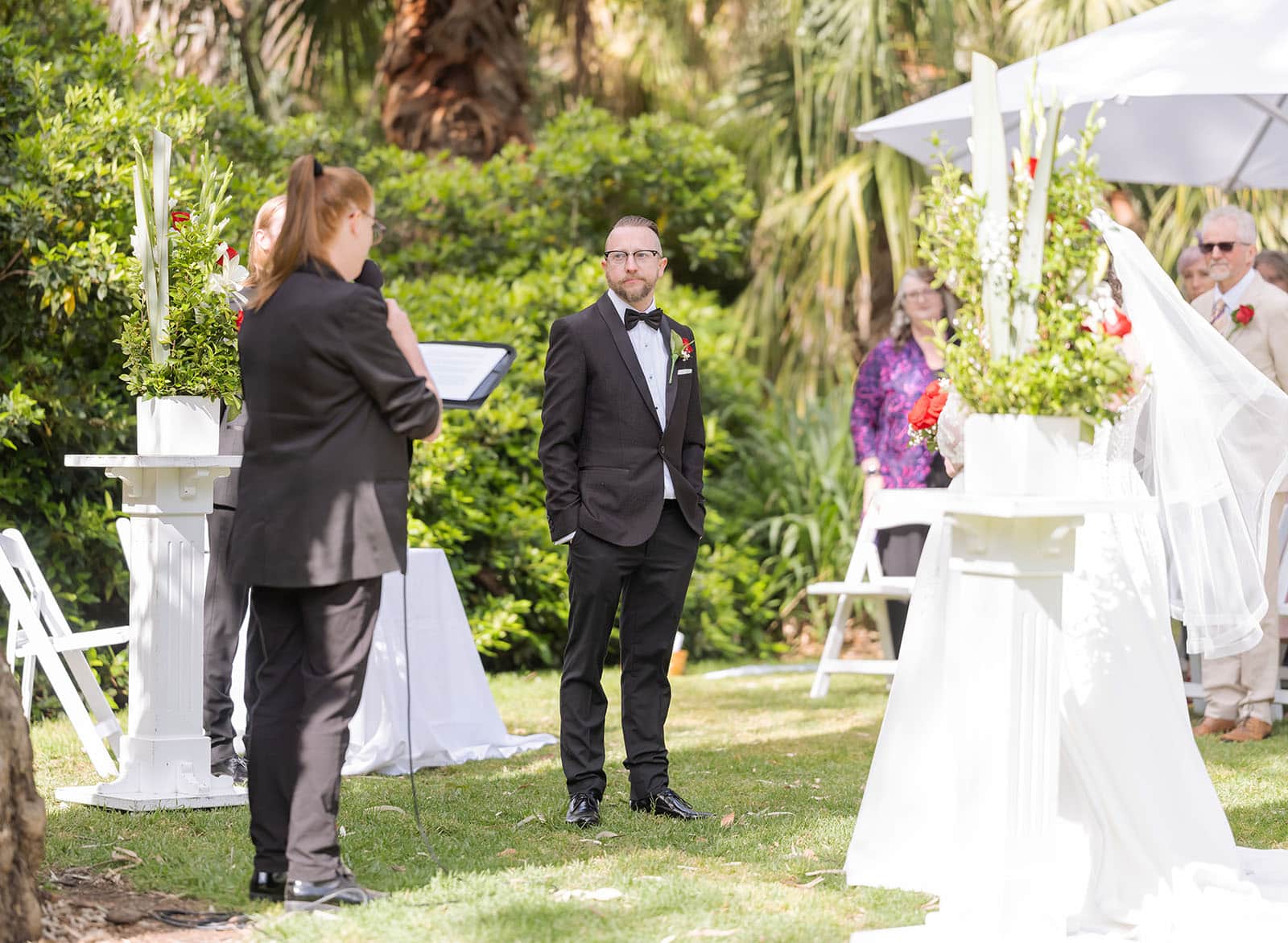 botanic gardens palm house wedding - kelly & david 96 Groom in a black tuxedo watching the bride approach during an outdoor garden wedding ceremony