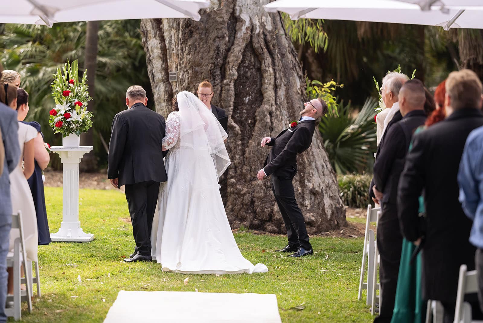 botanic gardens palm house wedding - kelly & david 93 Groom laughing during an outdoor wedding ceremony as the bride stands beside an officiant beneath a large gum tree