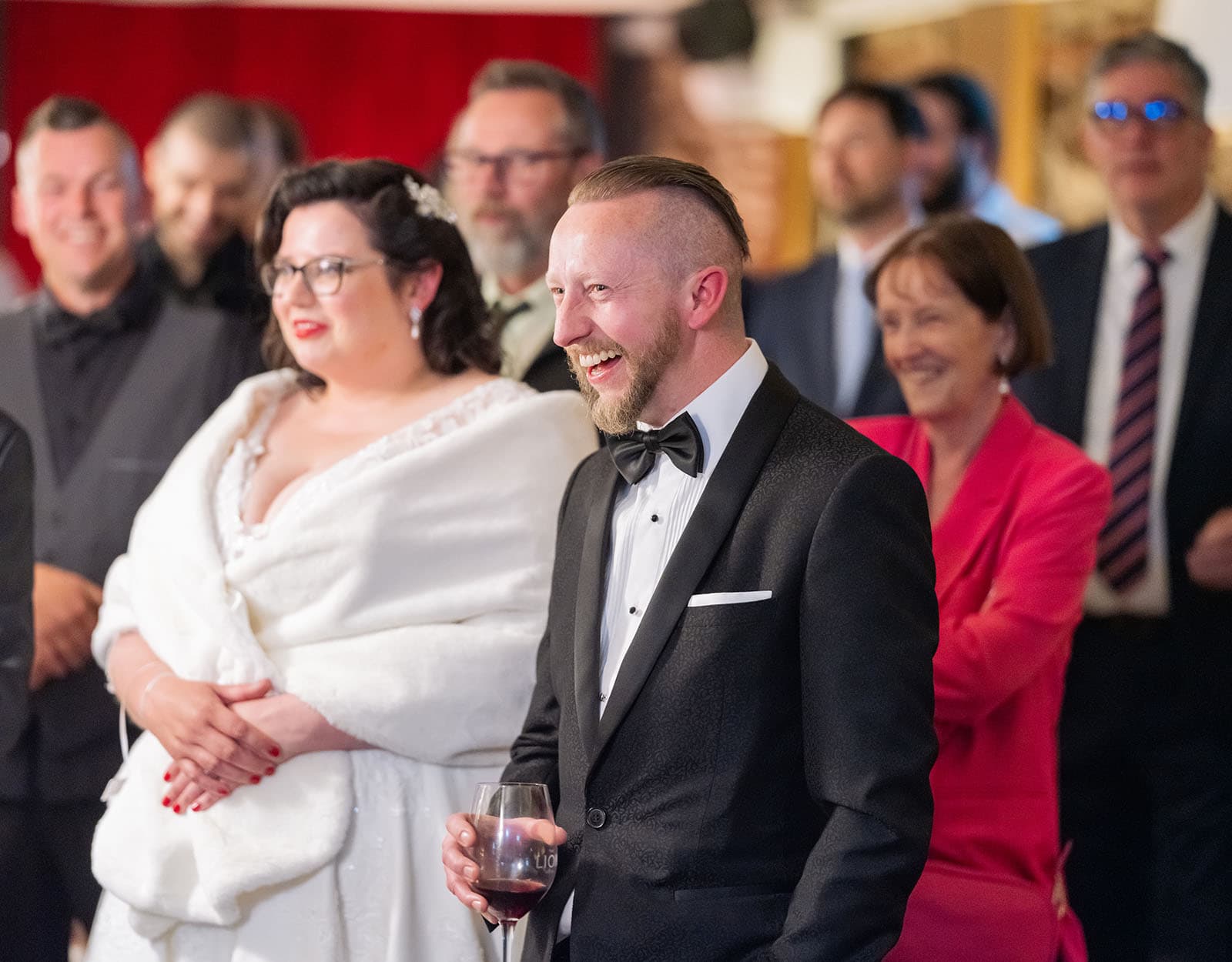 botanic gardens palm house wedding - kelly & david 131 Groom in a black tuxedo laughing while holding a glass of red wine beside the bride during a wedding speech