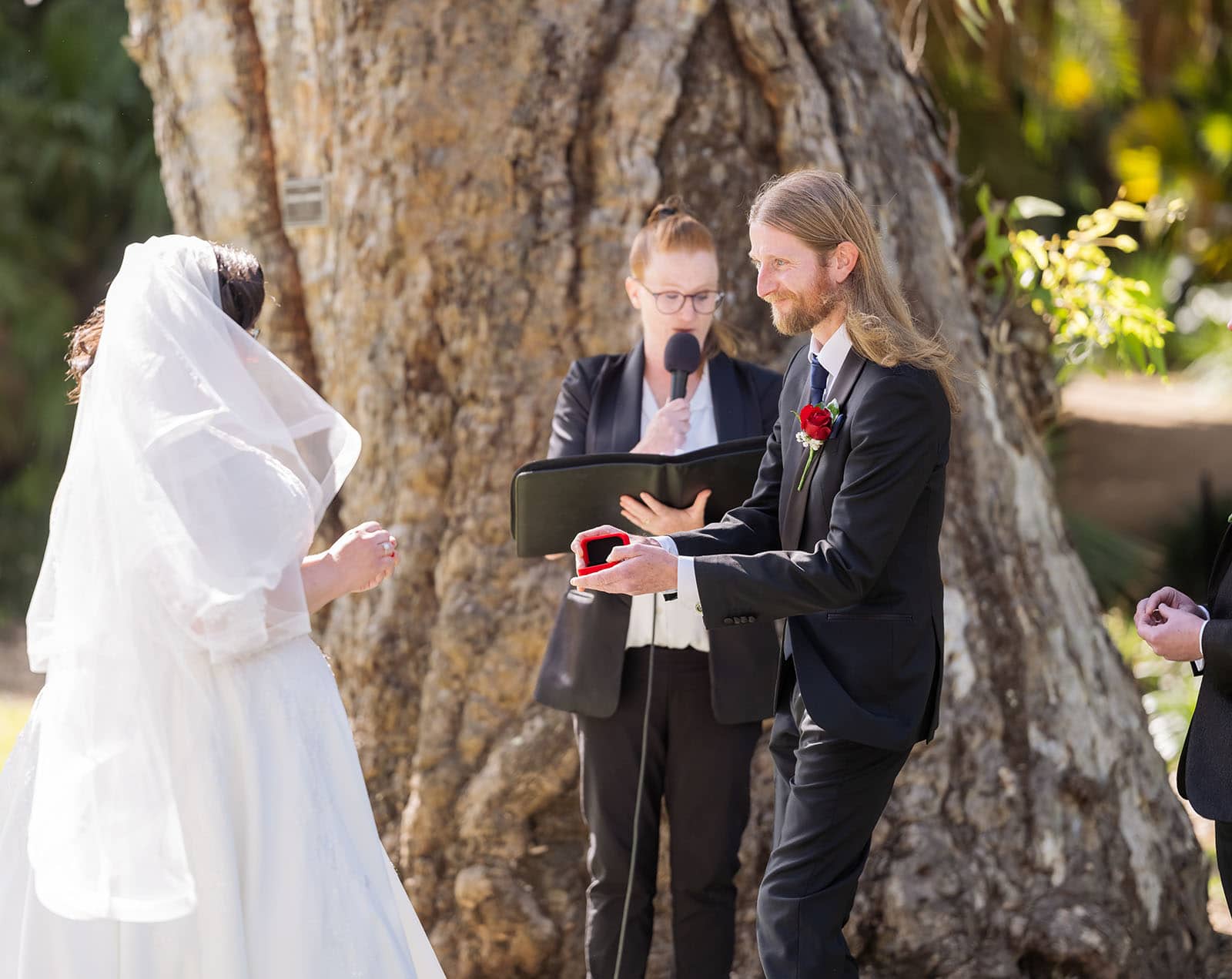 botanic gardens palm house wedding - kelly & david 99 Groom opens a red wedding ring box for his veiled bride during an outdoor ceremony, with the celebrant behind them.