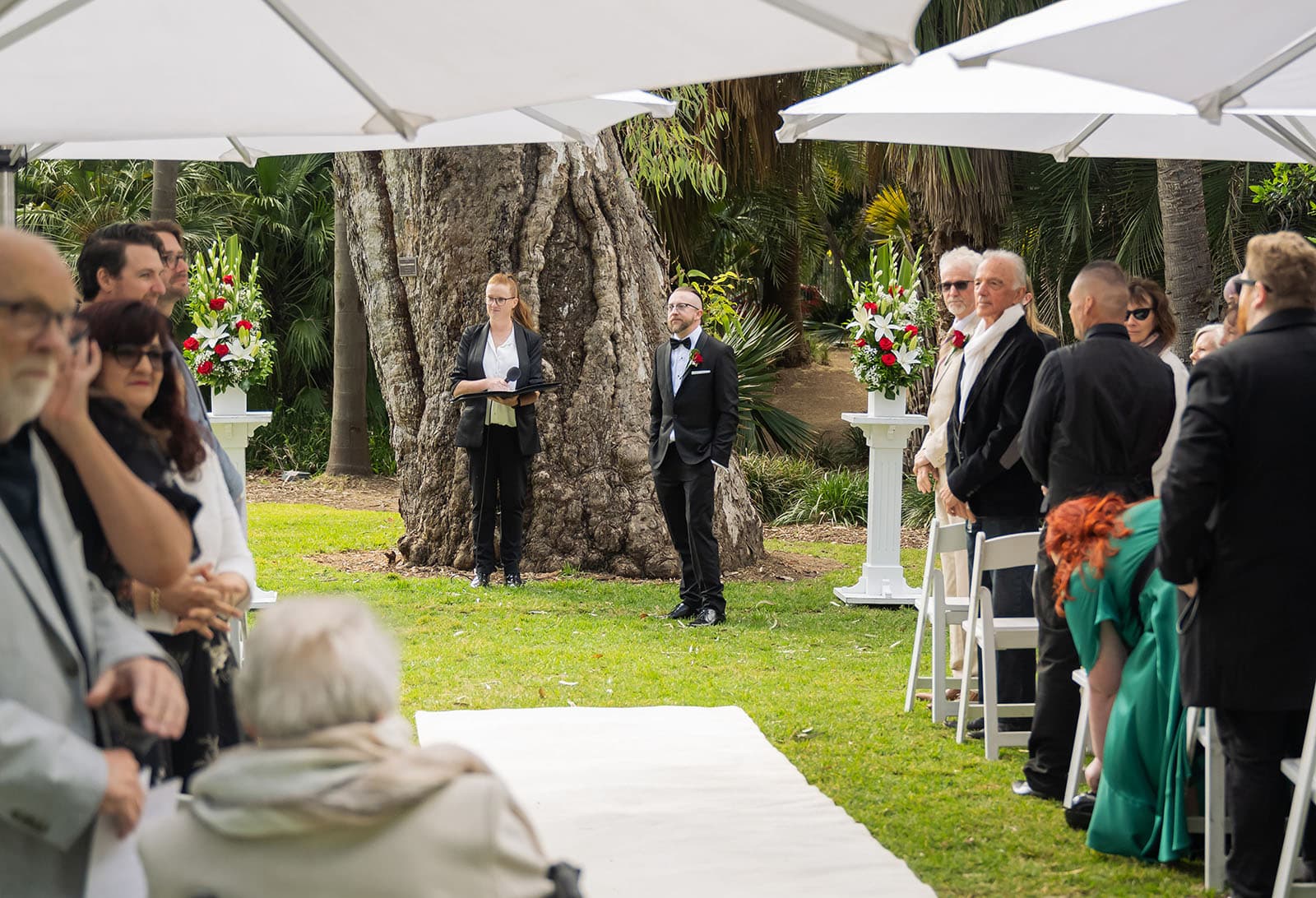 botanic gardens palm house wedding - kelly & david 91 Groom waits beside the celebrant at the end of a white aisle as wedding guests stand beneath umbrellas in Adelaide Botanic Gardens.