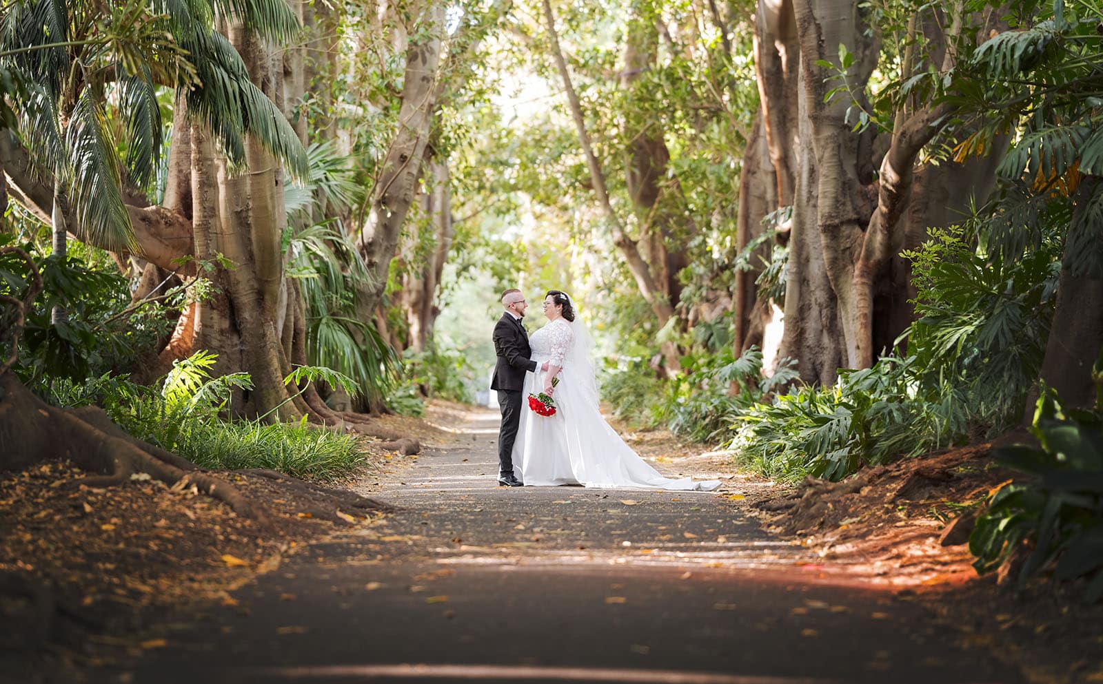 botanic gardens palm house wedding - kelly & david 111 Kelly and David standing together on a tree-lined path at Adelaide Botanic Gardens, with the bride holding a red bouquet.