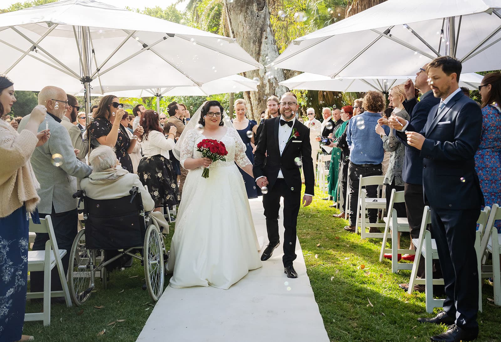 botanic gardens palm house wedding - kelly & david 104 Kelly and David walk down the garden wedding aisle as guests blow bubbles beneath white parasols at Adelaide Botanic Gardens.