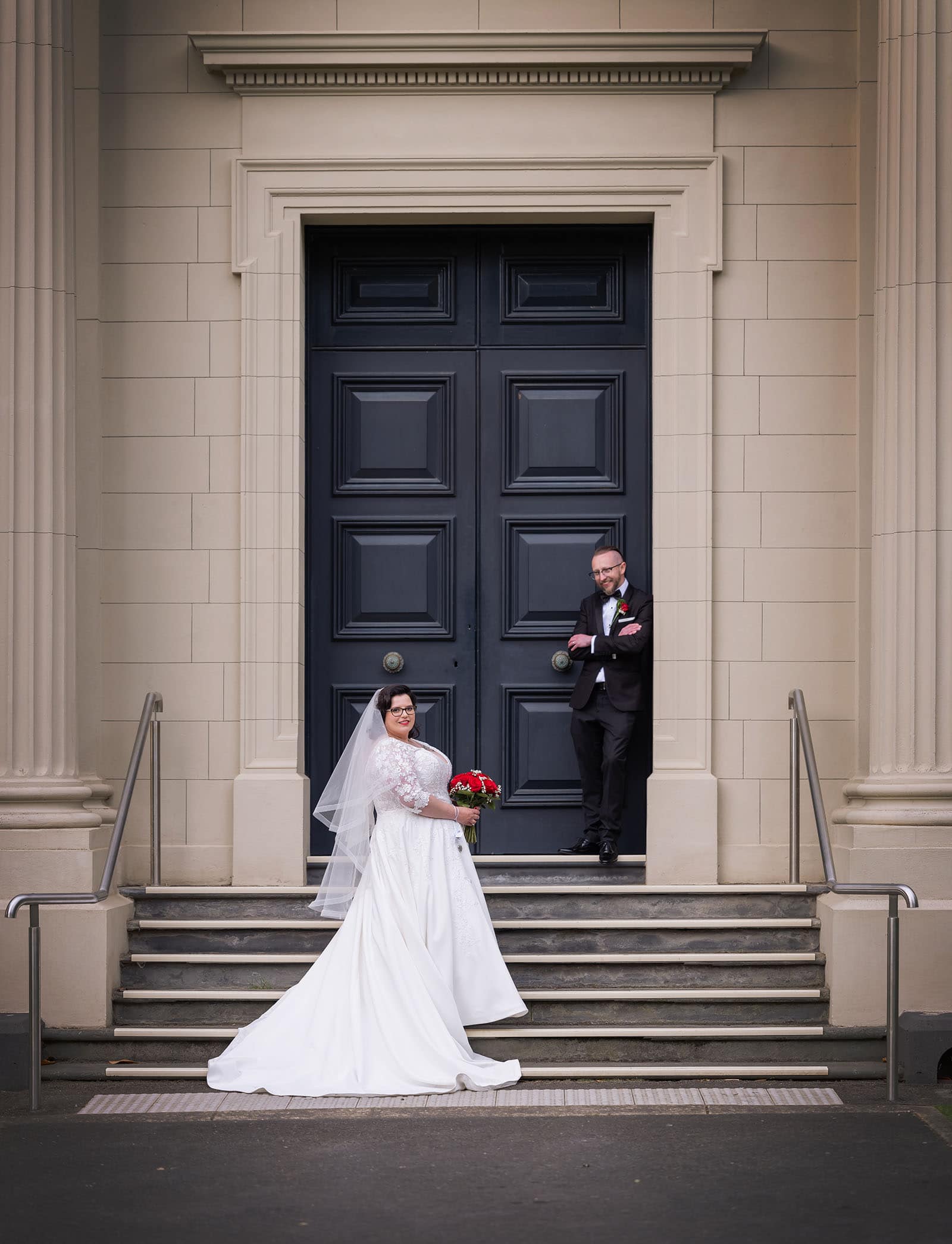 botanic gardens palm house wedding - kelly & david 117 Bride holding a red bouquet on stone steps as the groom stands in the doorway of the Economic Building