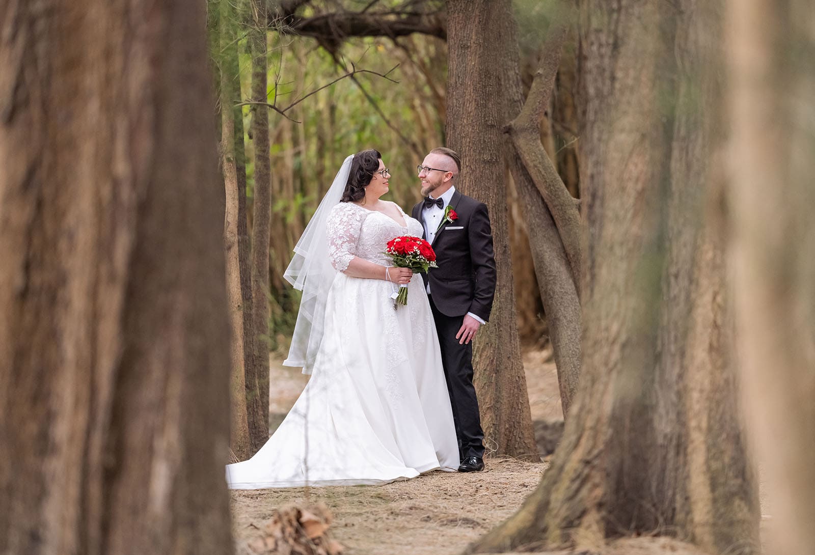 botanic gardens palm house wedding - kelly & david 116 Kelly and David smiling at one another between tree trunks, with Kelly holding a red bouquet in her white wedding dress and David wearing a black tuxedo.