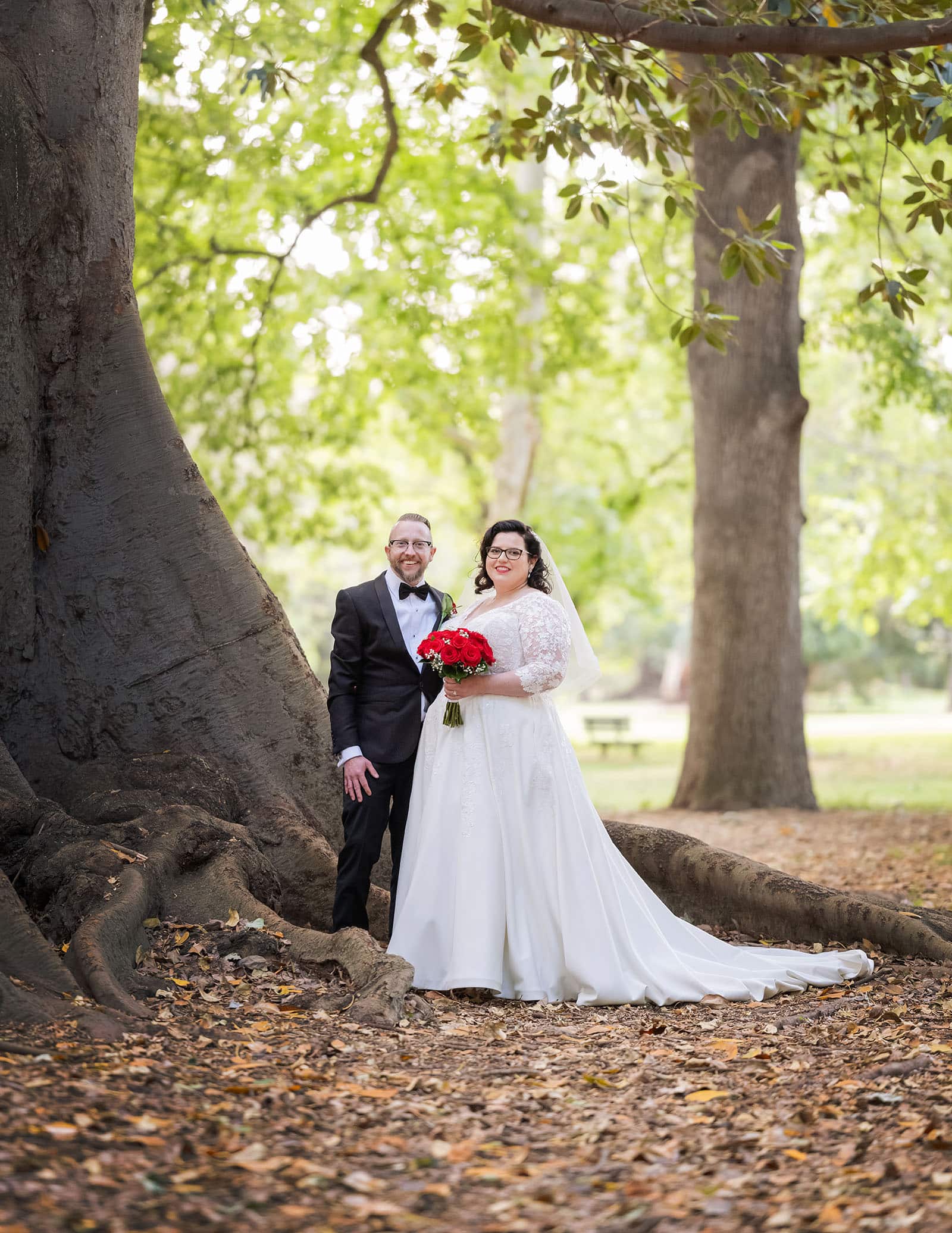 botanic gardens palm house wedding - kelly & david 119 Kelly and David stand beside the sprawling roots of a Moreton Bay fig tree, with the bride holding a red rose bouquet at Adelaide Botanic Gardens.