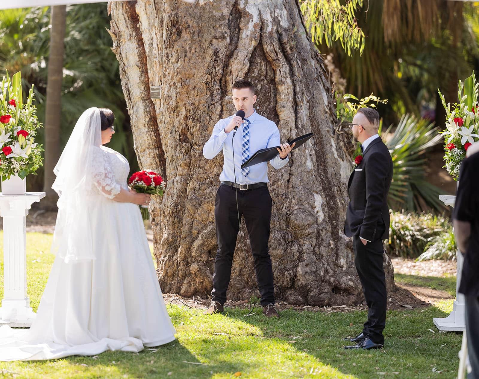 botanic gardens palm house wedding - kelly & david 98 Bride and groom listen as a man speaks into a microphone beneath a large gum tree during their Adelaide Botanic Gardens ceremony.