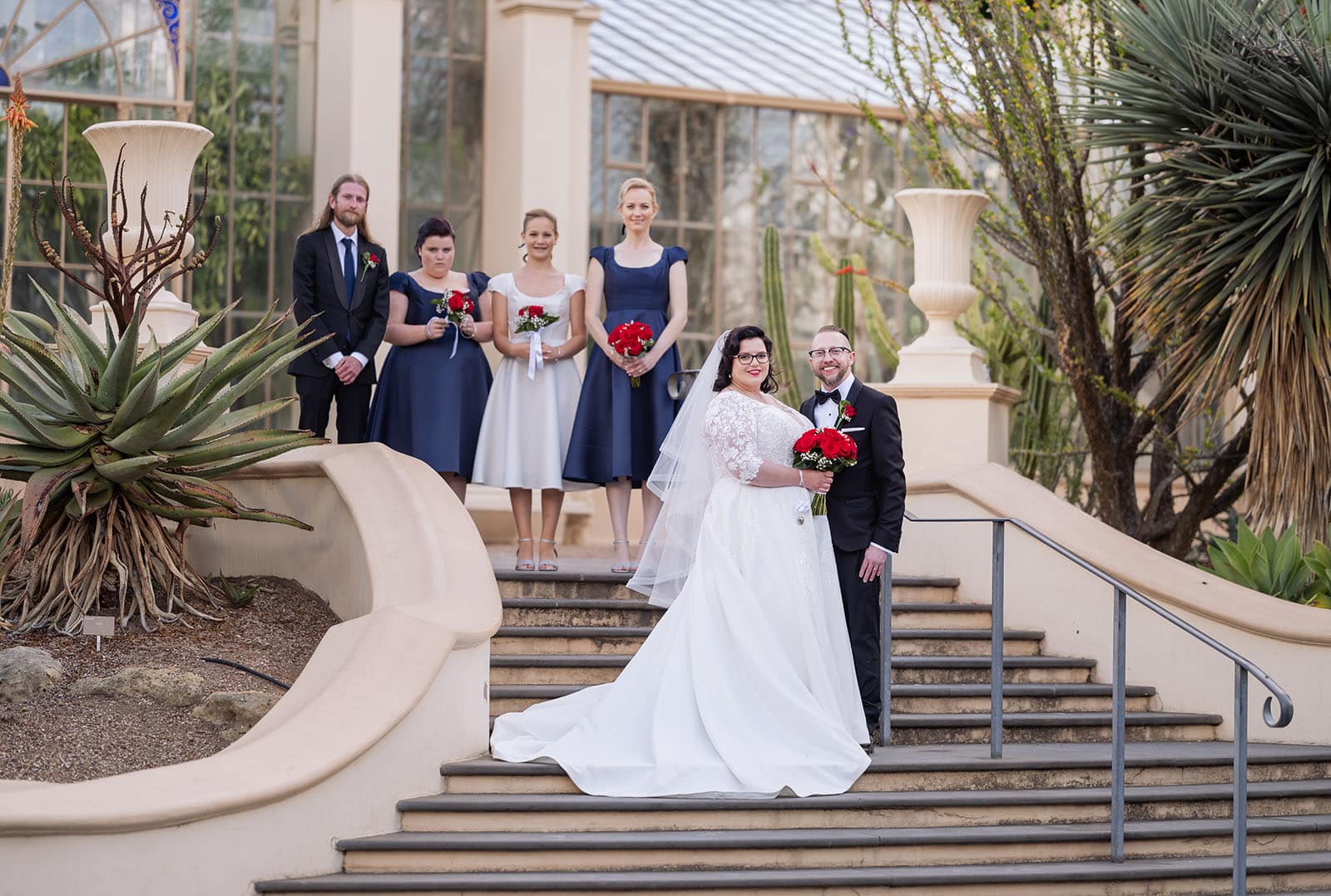 botanic gardens palm house wedding - kelly & david 113 Bride and groom holding red bouquets with their wedding party on steps outside the Palm House at Adelaide Botanic Gardens