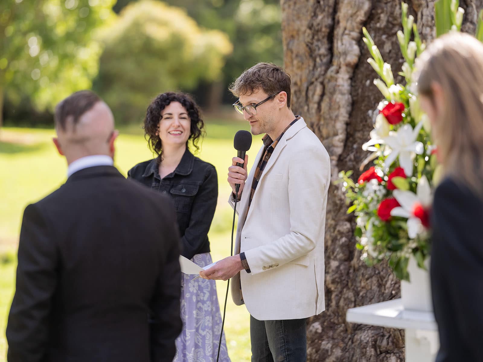 botanic gardens palm house wedding - kelly & david 97 Man in a light jacket reading into a microphone during an outdoor wedding ceremony beside red and white flowers