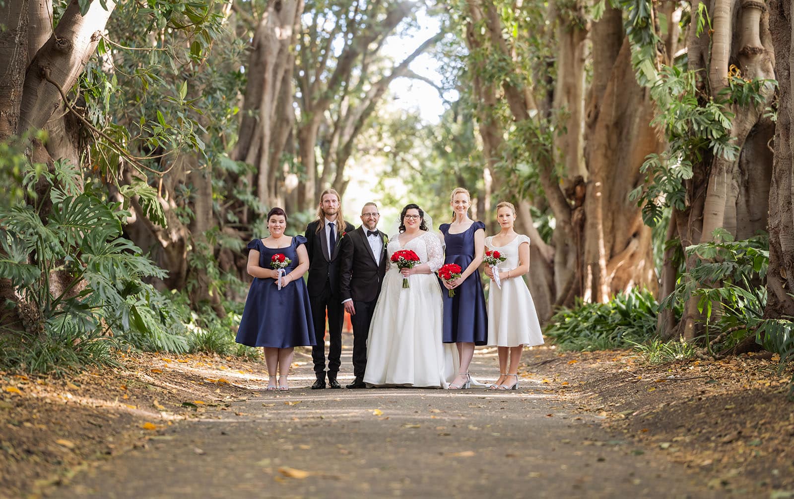 botanic gardens palm house wedding - kelly & david 110 Kelly and David with their wedding party holding red bouquets on a tree-lined path at Adelaide Botanic Gardens