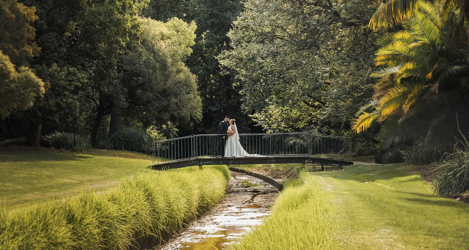 botanic gardens, araucaria avenue wedding - jade & hazim 46 Romantic couple embracing on a quaint bridge over a stream in a lush, sunlit garden.