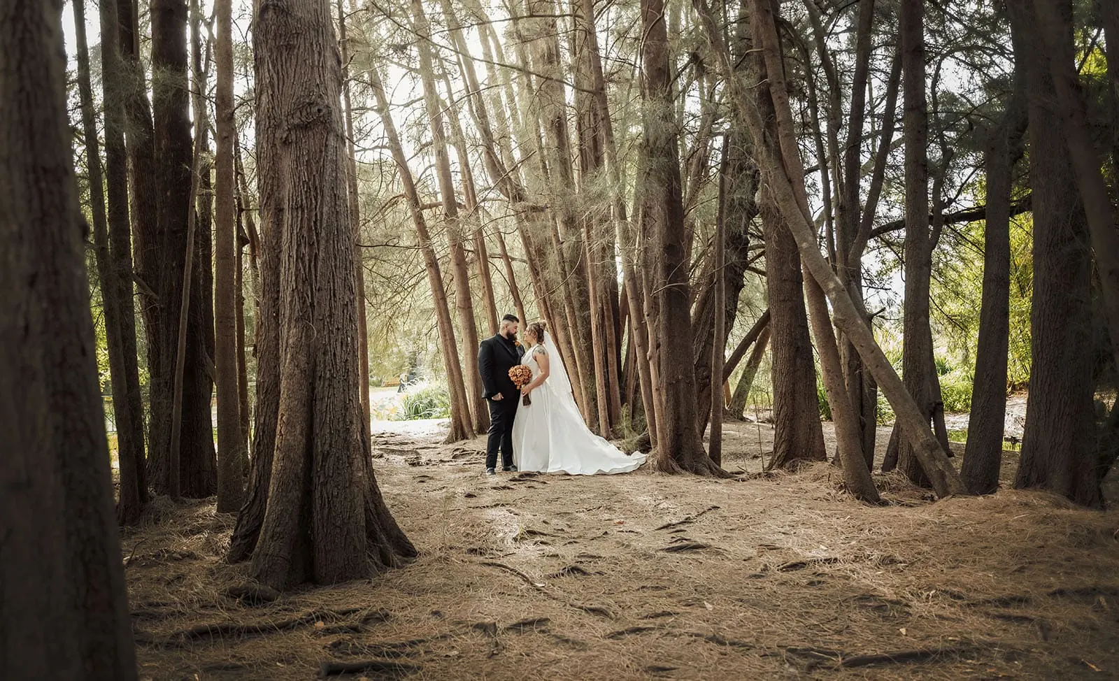 botanic gardens, araucaria avenue wedding - jade & hazim 57 Couple embracing in wedding attire amidst serene pine forest.
