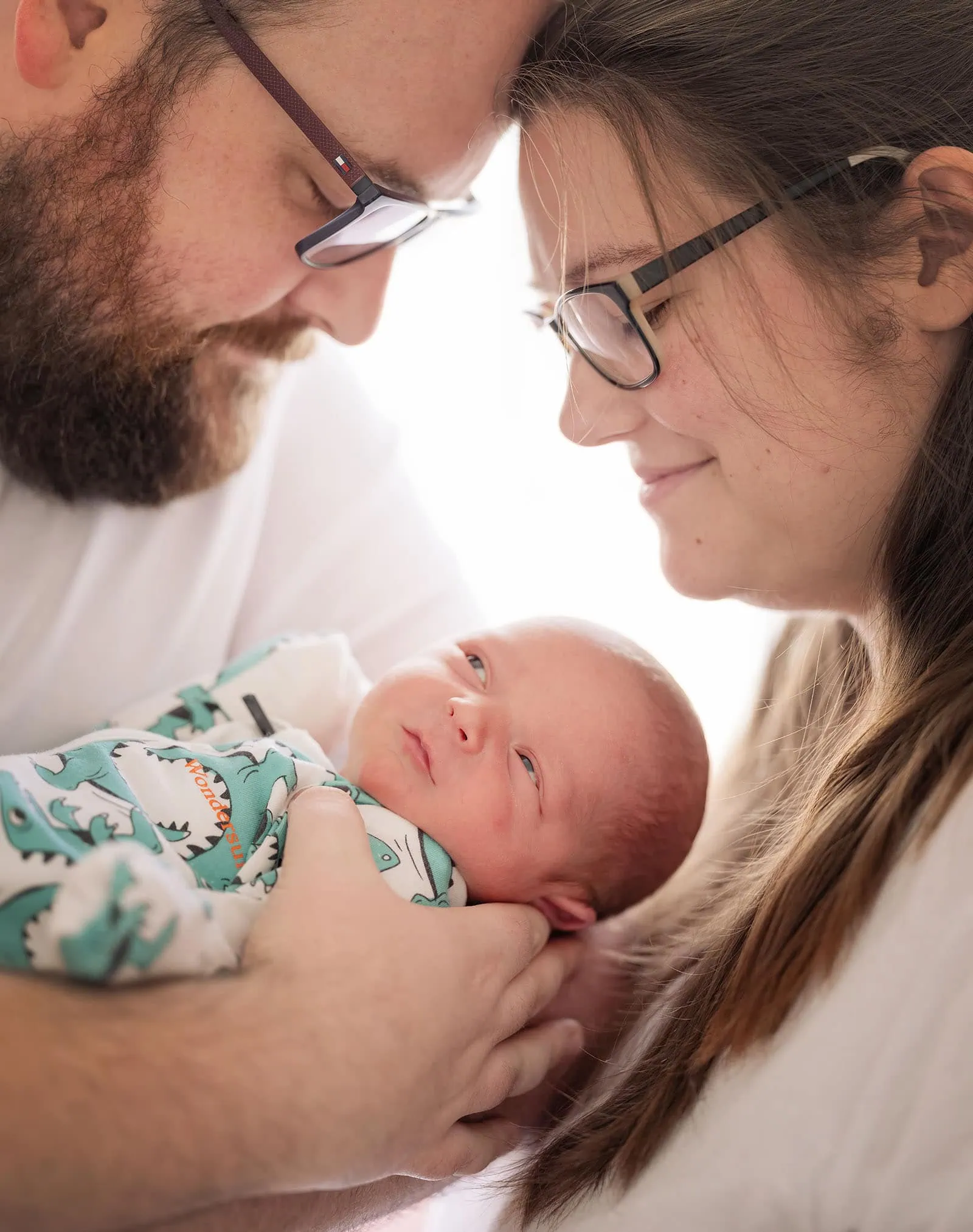 baby spencer's newborn shoot 65 Parents in glasses lovingly bonding with their newborn baby dressed in a dinosaur onesie.