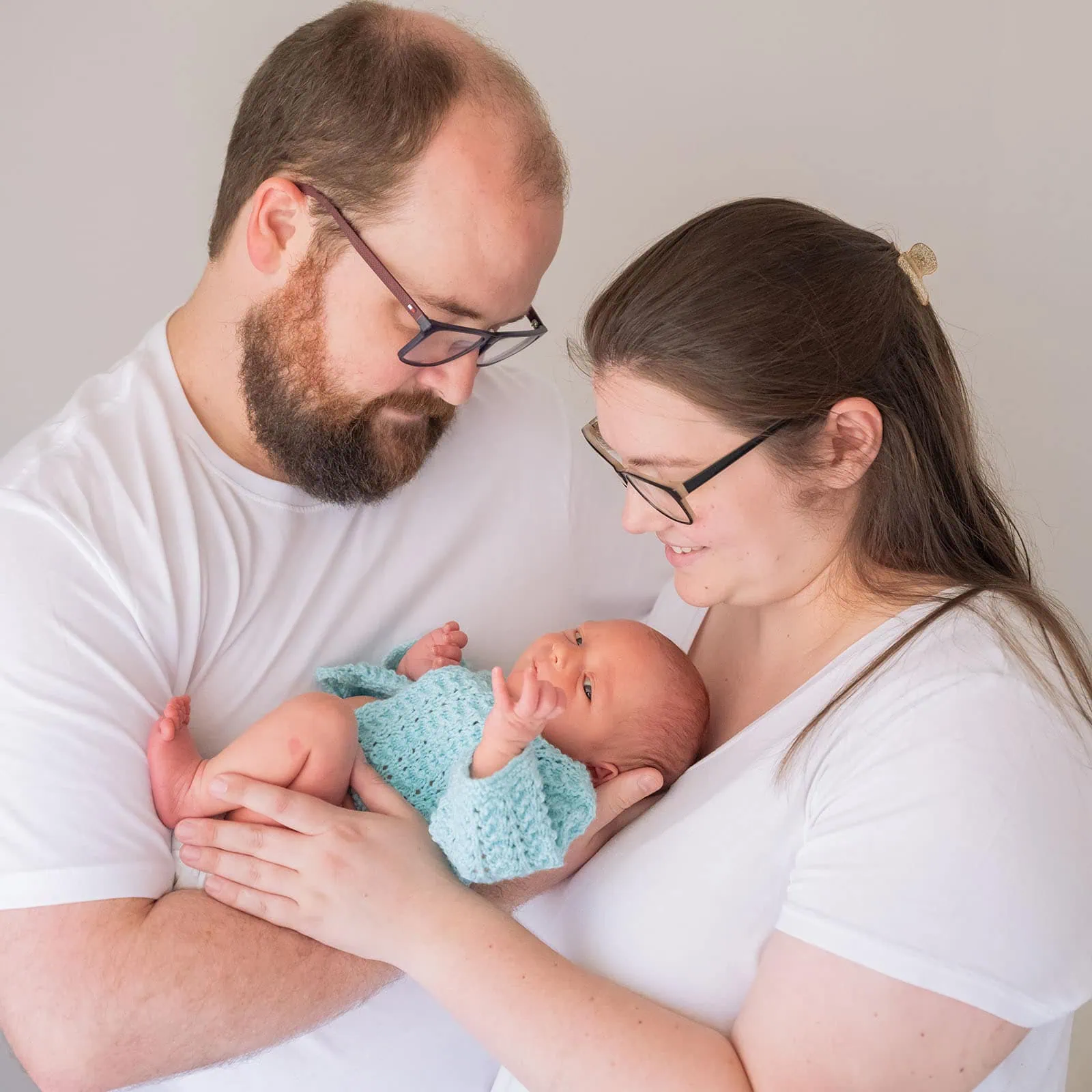 baby spencer's newborn shoot 79 Parents in white shirts adoring their newborn baby in a blue knitted garment.