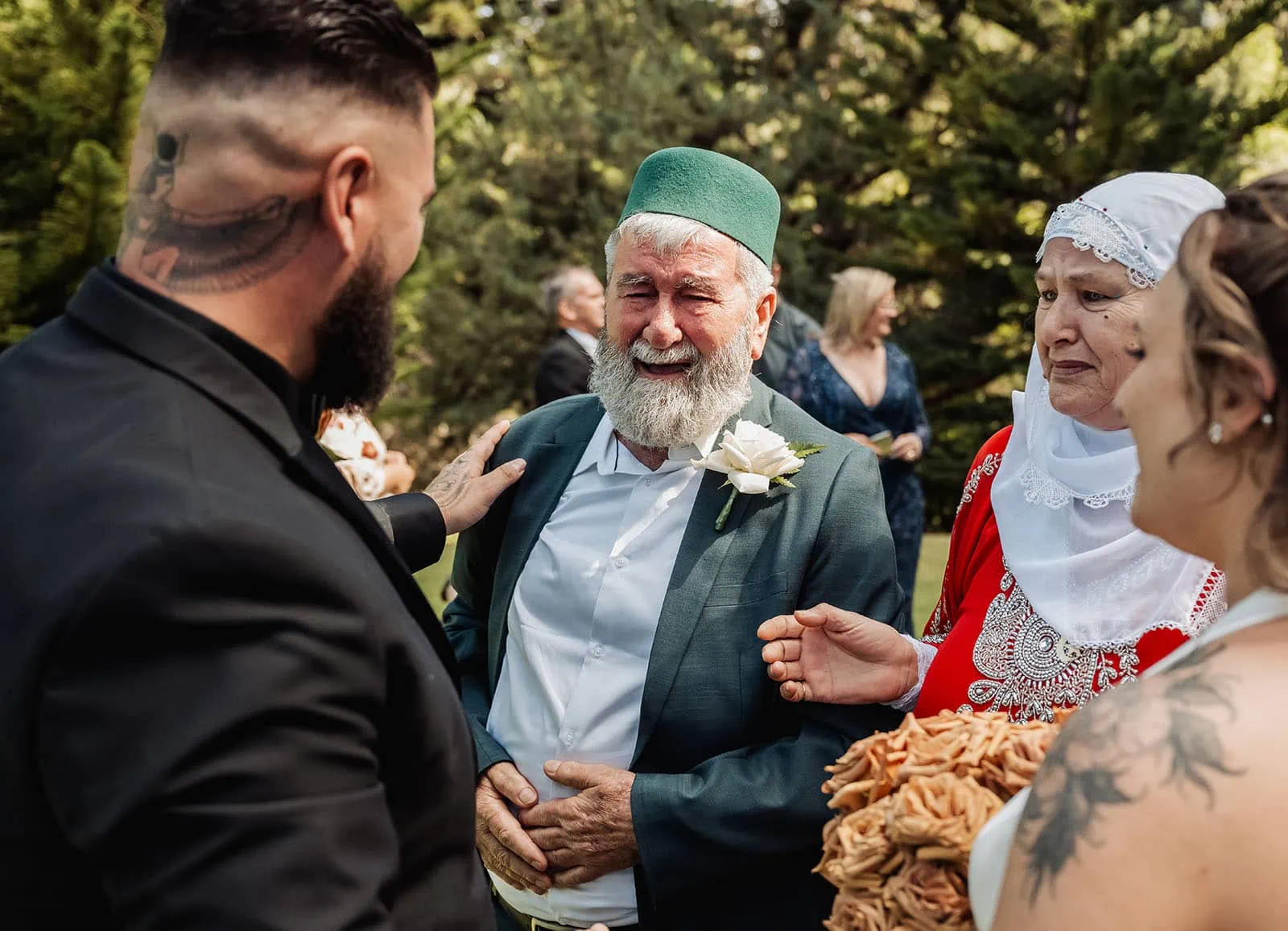 botanic gardens, araucaria avenue wedding - jade & hazim 42 Elderly man sharing emotional moment at outdoor wedding with tattooed guest and women in traditional attire.