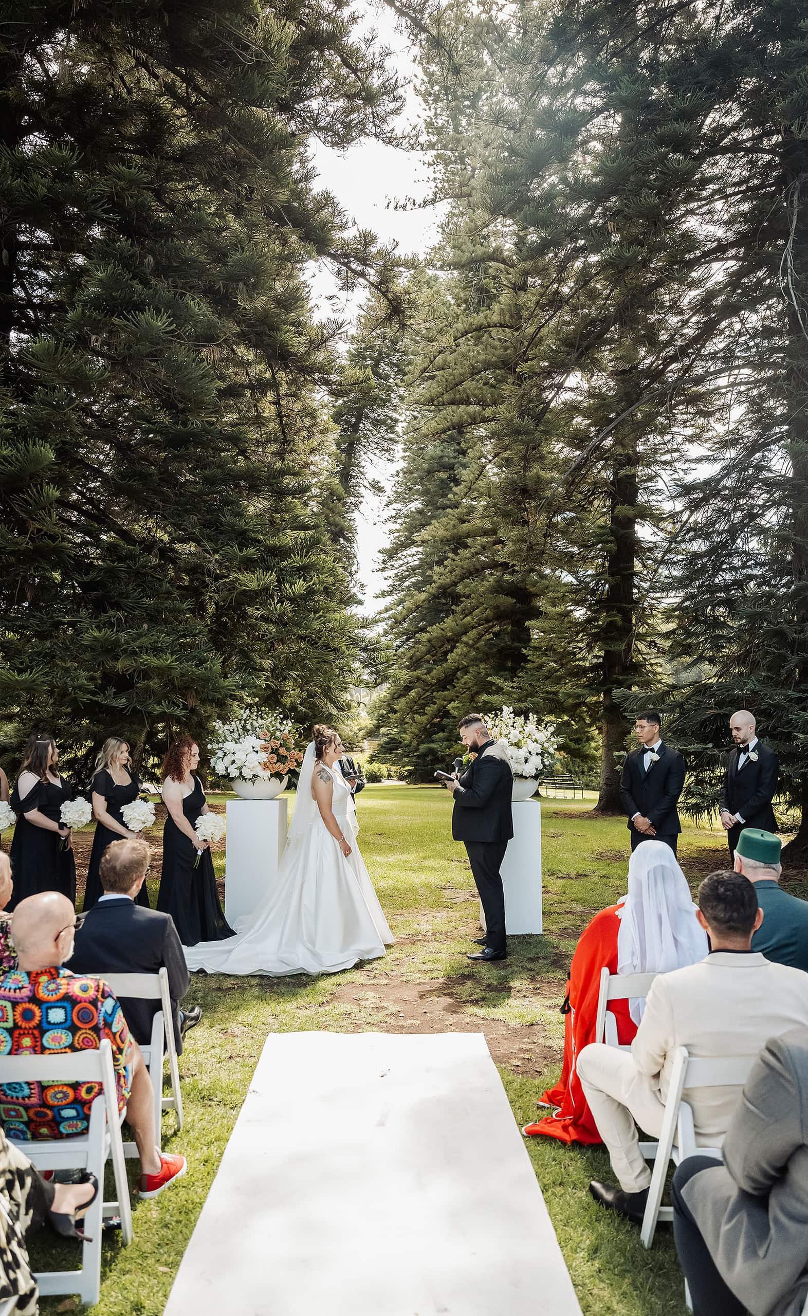 botanic gardens, araucaria avenue wedding - jade & hazim 36 Bride and groom exchanging vows at outdoor wedding amidst towering coniferous trees.