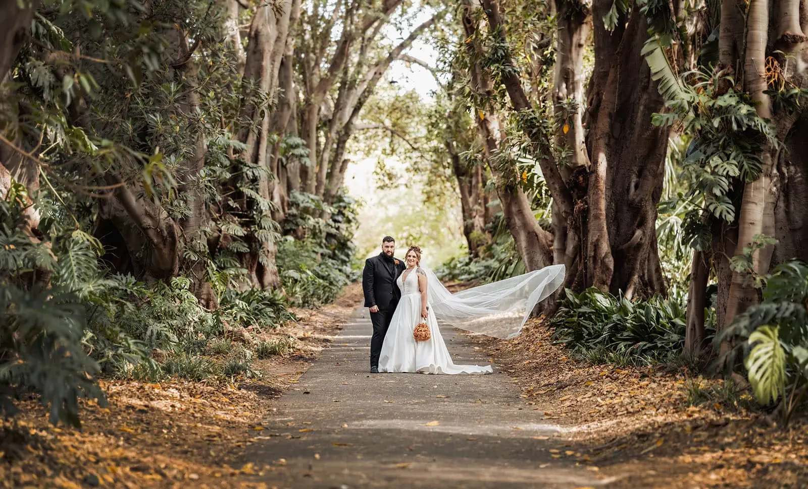 botanic gardens, araucaria avenue wedding - jade & hazim 51 Joyful couple celebrating autumn wedding under tree canopy in serene park.
