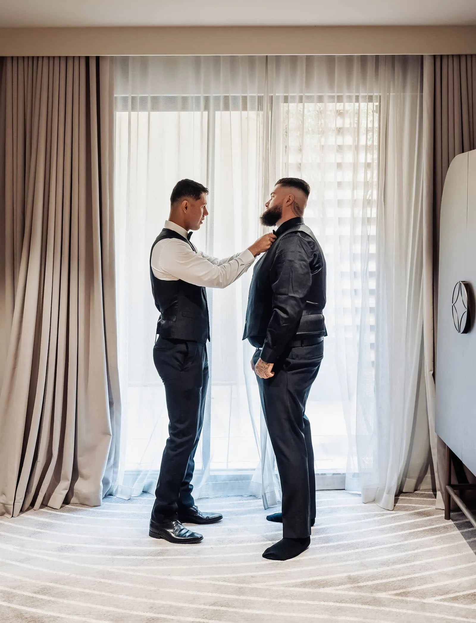 botanic gardens, araucaria avenue wedding - jade & hazim 4 Two stylish men adjusting attire in a sunlit, elegant room before a formal event.