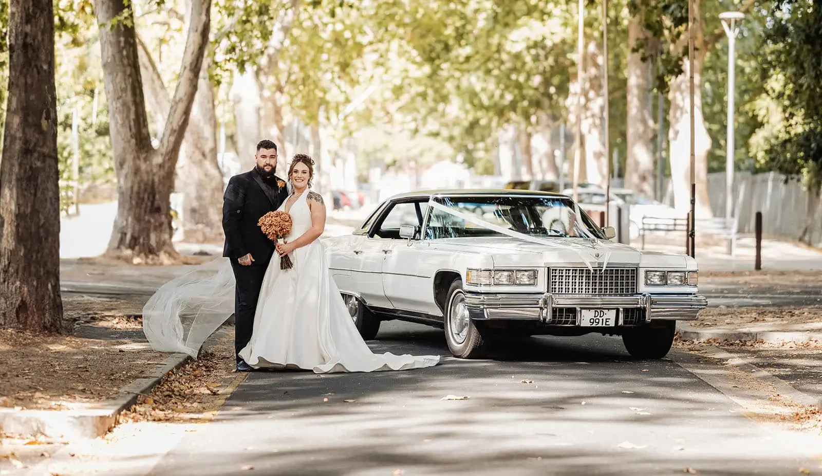 botanic gardens, araucaria avenue wedding - jade & hazim 44 Newlywed couple posing joyously beside a classic Cadillac on a tree-lined street.