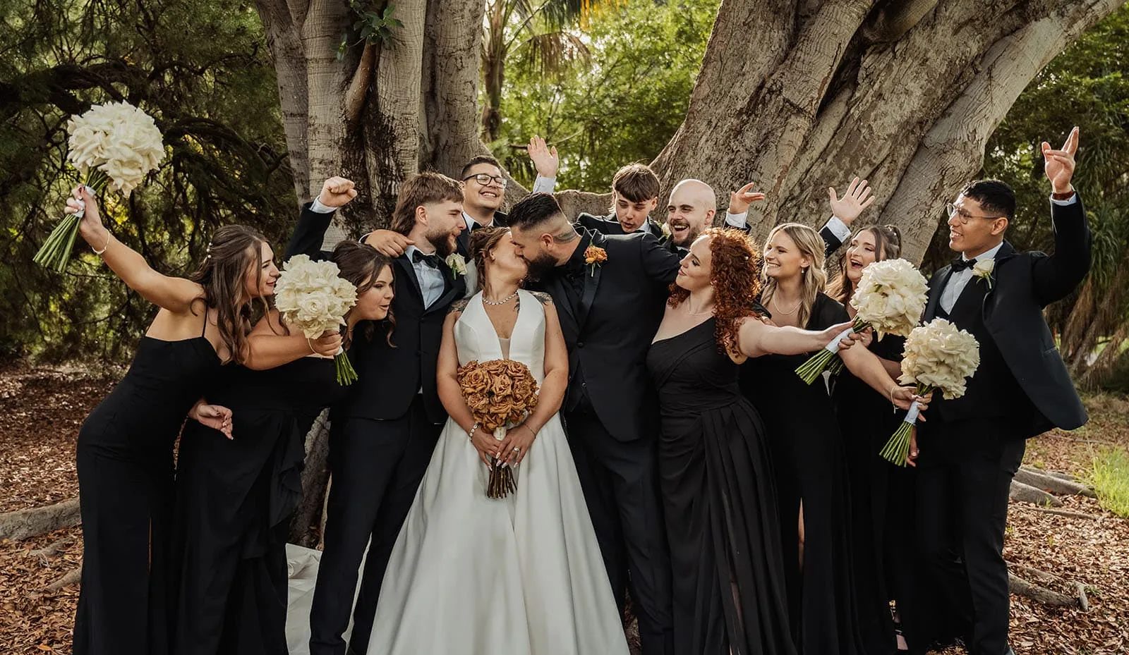 botanic gardens, araucaria avenue wedding - jade & hazim 56 Newlywed couple kissing amidst celebrating bridal party in an outdoor setting.