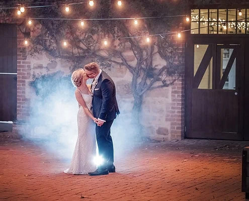 Couple sharing intimate moment in dreamy wedding setting with string lights and fog.