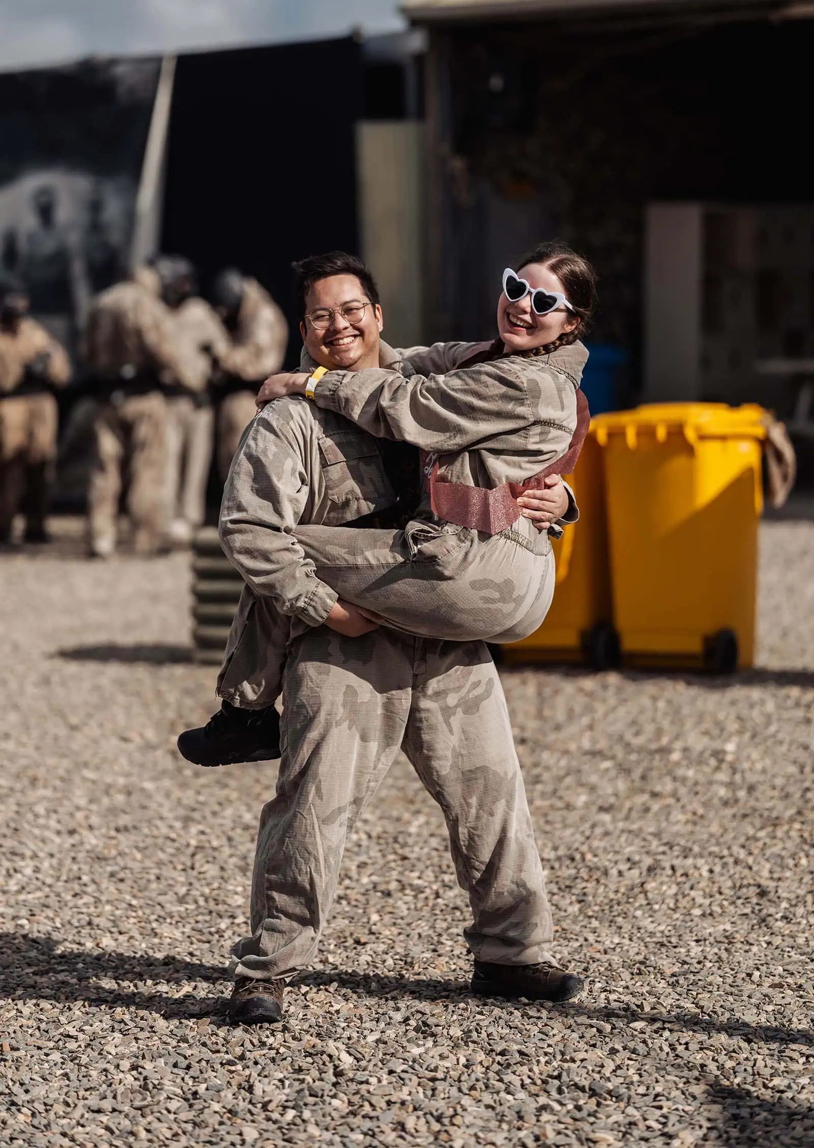 monarto delta force bucks vs hens paintball party 6 Two friends in camouflage jumpsuits, one wearing heart-shaped sunglasses, share a playful moment.