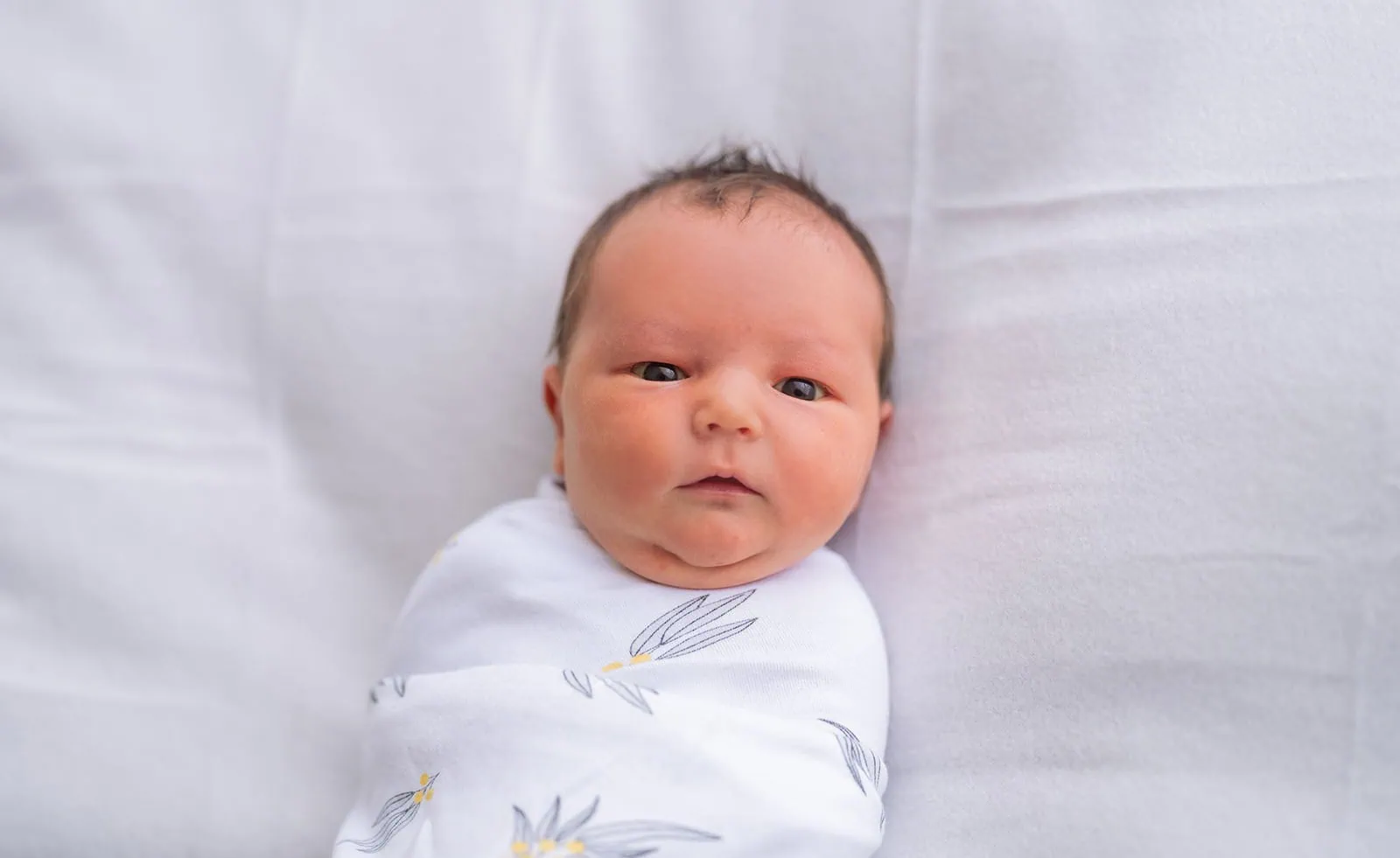 baby joshua's newborn photo shoot 29 Newborn baby swaddled in white botanical blanket on white bed, looking at camera.