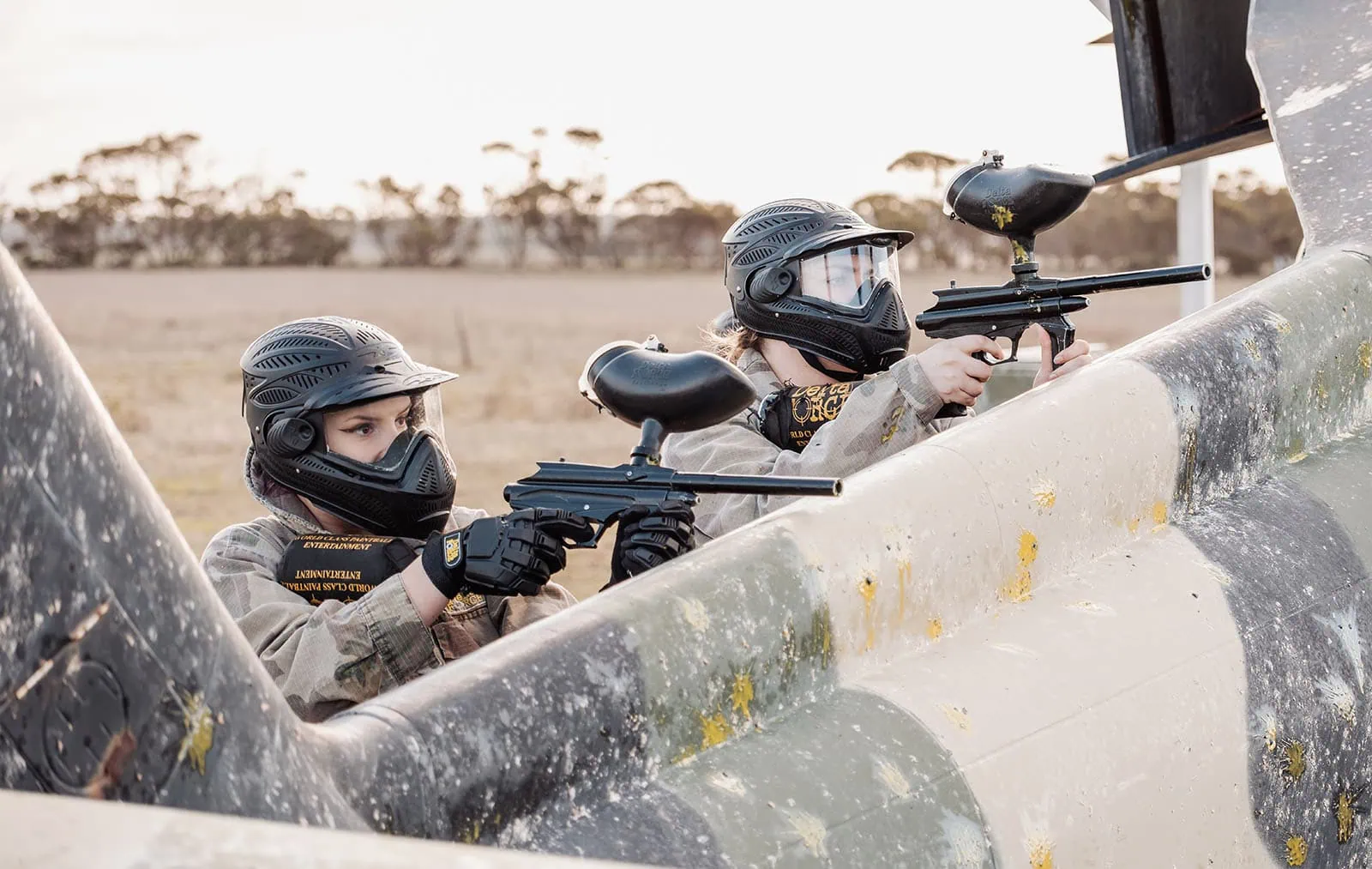monarto delta force bucks vs hens paintball party 34 Paintball players in action behind a camouflaged barricade, aiming paintball guns.