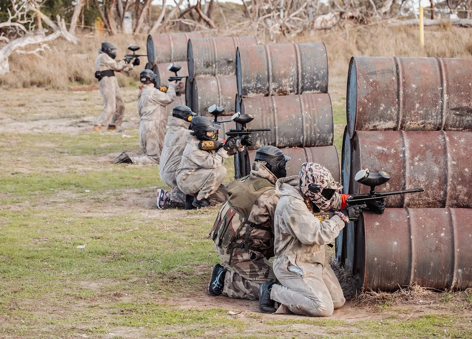 monarto delta force bucks vs hens paintball party 31 Paintball players in camouflage gear taking cover behind barrels in an outdoor field.