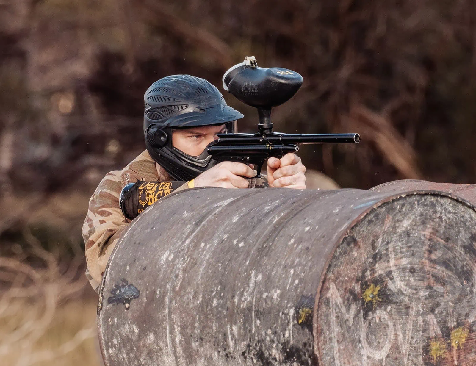 monarto delta force bucks vs hens paintball party 27 Paintball player aiming marker behind barrel in wooded area.