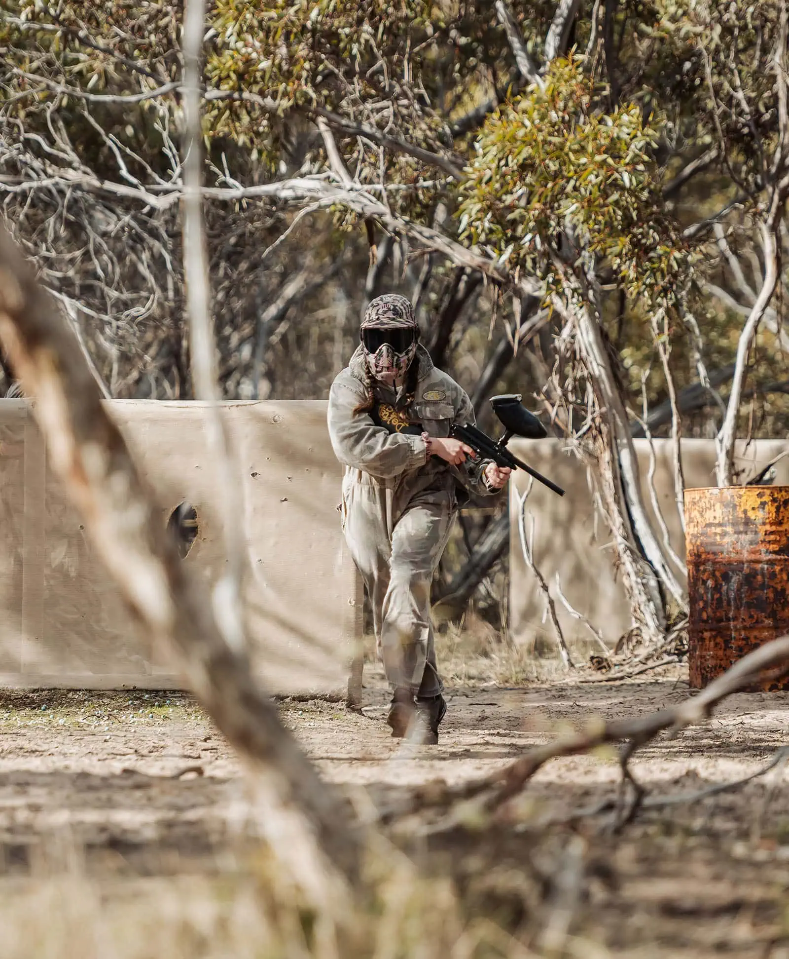 monarto delta force bucks vs hens paintball party 15 Paintball player in camouflage aiming gun in wooded area with barricade and barrel.