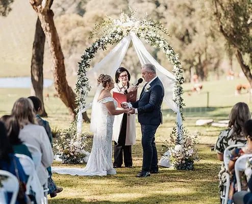 Outdoor wedding ceremony with a couple under a decorated arch, surrounded by nature and guests.