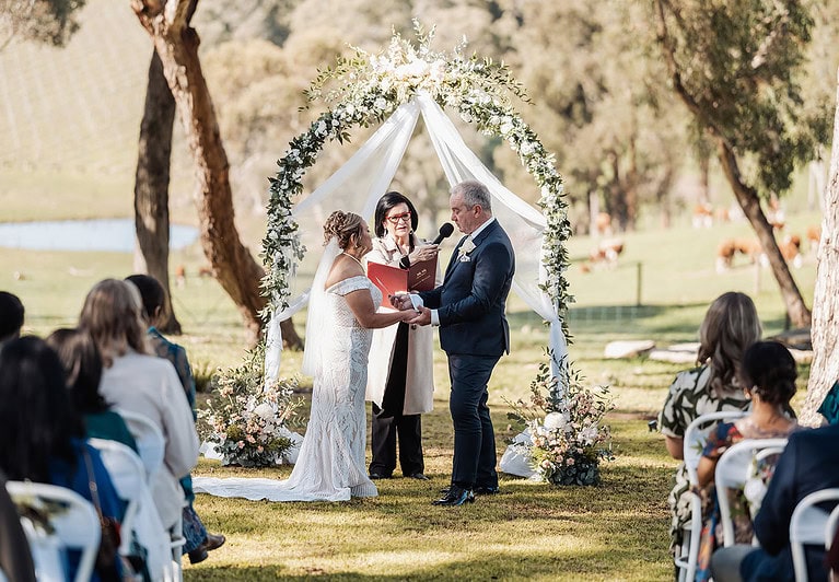 Outdoor wedding ceremony with a couple under a decorated arch, surrounded by nature and guests.