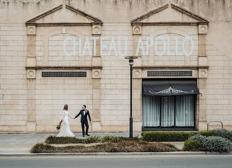 Newlyweds at Chateau Apollo in wedding attire, vintage architecture background.