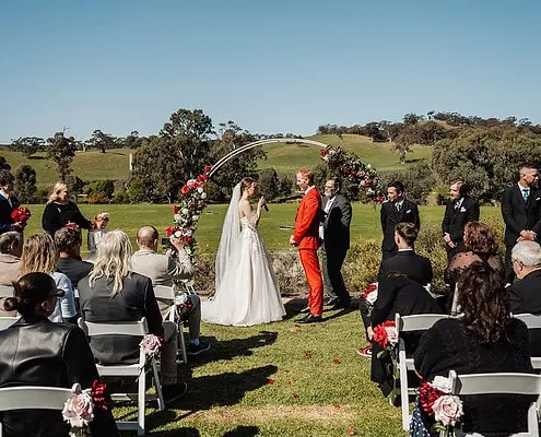 Outdoor wedding with floral arch, bride and groom, guests on lawn under blue sky.