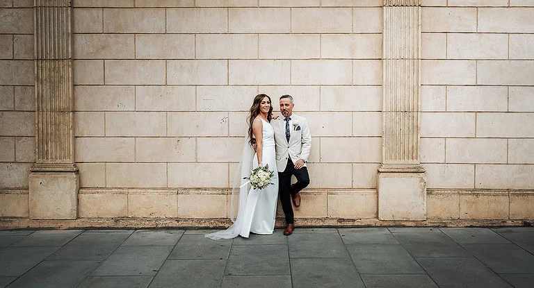 Elegant newlywed couple poses against textured stone wall, wearing coordinated outfits, exuding timeless elegance.