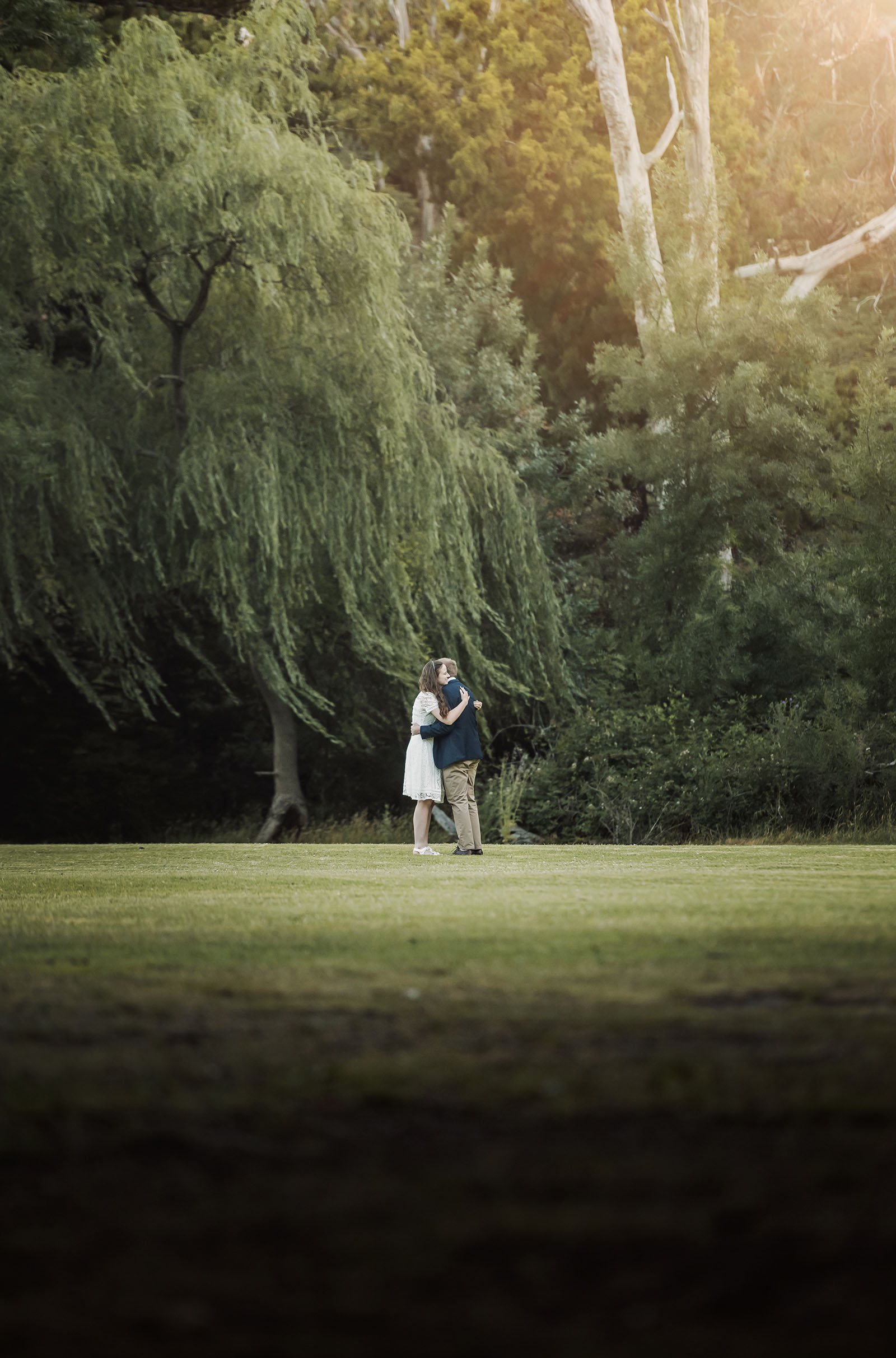 romantic belair national park proposal - marissa & james 137 Marissa and James embracing on a grassy lawn beneath willow trees at Belair National Park