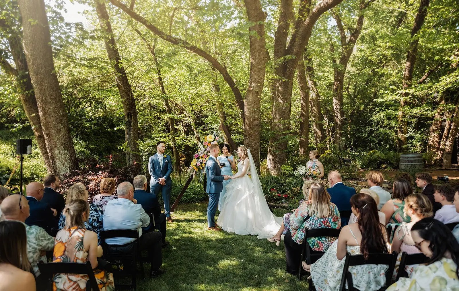 inglewood inn 10 Lush forest wedding ceremony with bride, groom, floral arch, guests, and vibrant greenery.