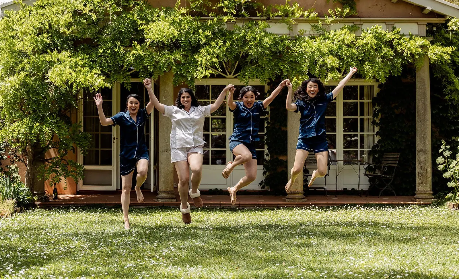 the night before the wedding: how brides can wake up with camera-ready hair 106 Women in pajamas jumping joyfully in lush garden with rustic cottage backdrop.