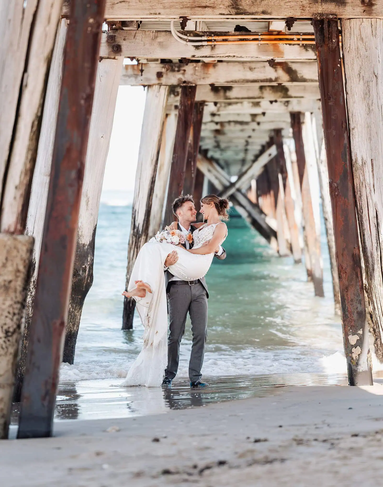 pt noarlunga underwater wreck the dress shoot - jess & jack 2 Bride and groom under rustic pier at beach, embracing love and serenity.