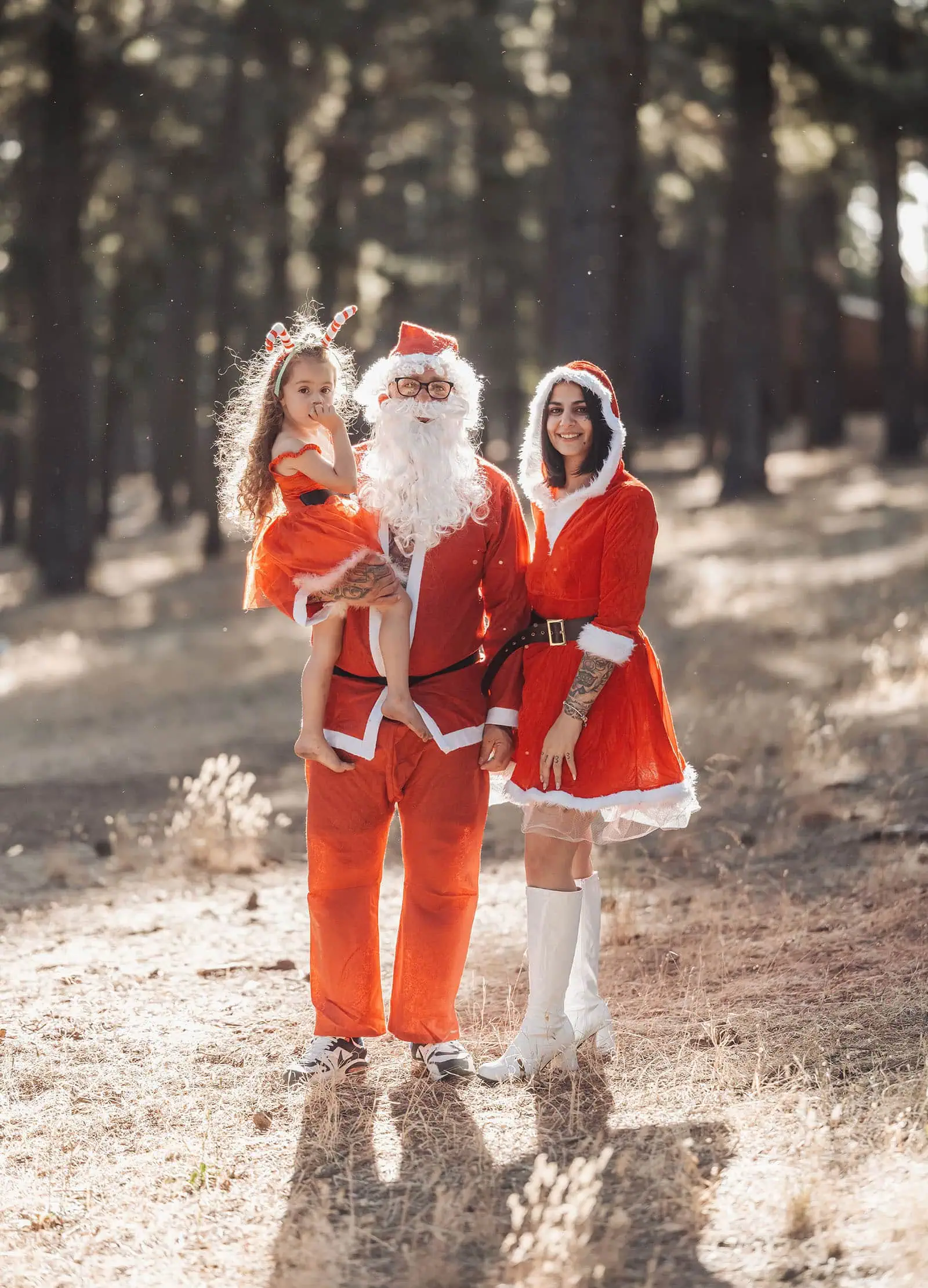 last minute father christmas photoshoot 42 Festive Santa, Mrs. Claus, and child in Christmas attire pose in a sunlit forest.