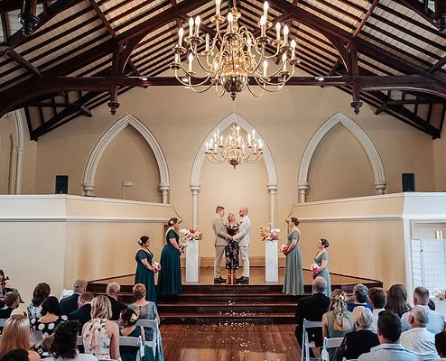 Wedding ceremony in ornate hall with floral arrangements, chandeliers, and elegantly dressed couple.