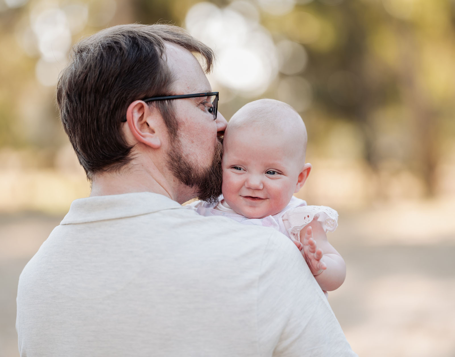 carisbrooke park family photoshoot 74 Man gently kisses baby’s forehead in sunlit outdoor setting, expressing love and warmth.